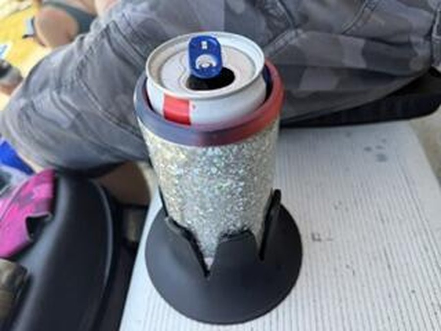 A black Steadi on a grandstand bench holding a canned drink in a glitter koozie, a spectator's leg beside it, at an outdoor race