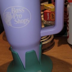 A lavender Bass Pro Shops insulated mug with a handle seated in a green drink base marked STEADI, standing on a cluttered wooden side table with jars and a red cup behind